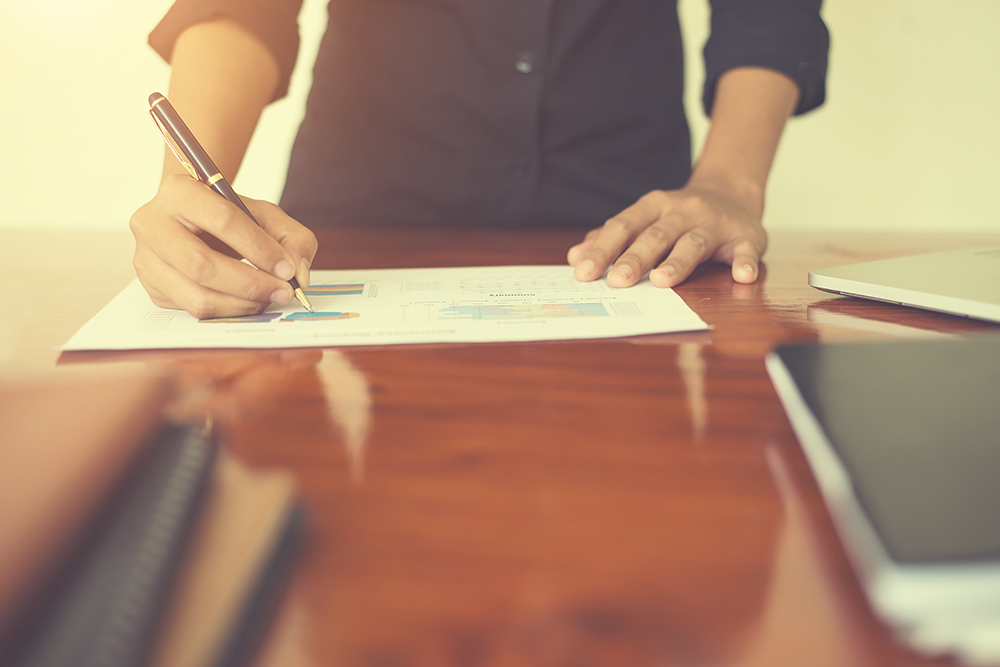 Person writing on a business report with charts on a wooden desk next to a laptop
